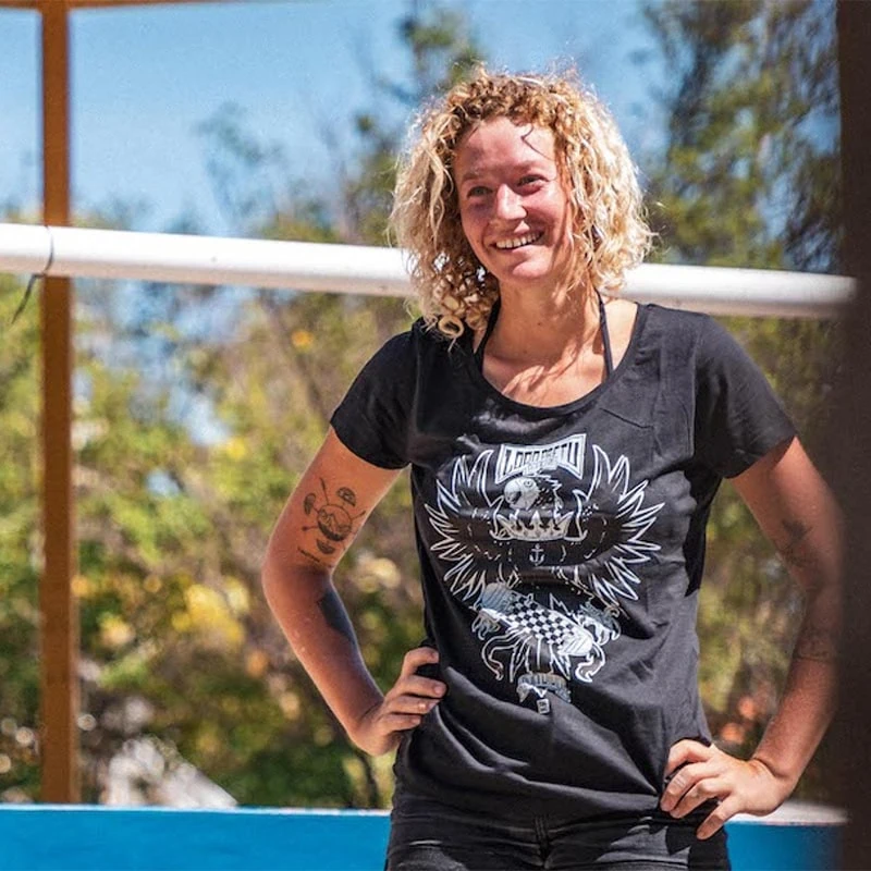 Blonde lady wears a black Swakiko ladies shirt with the design of the black parrot with the crown around his neck and a skateboard between his legs