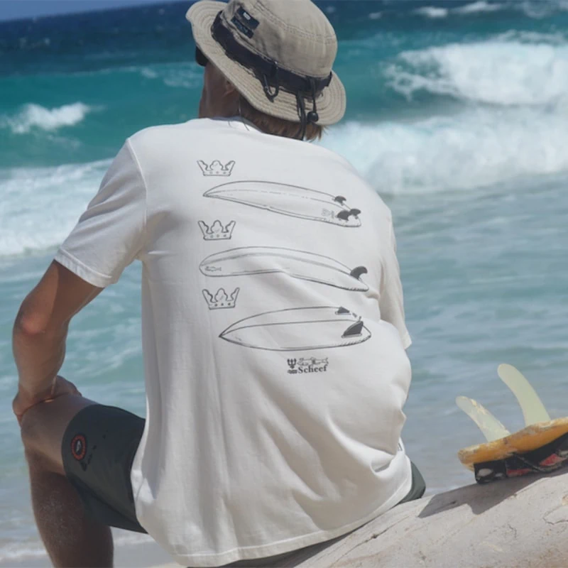 Model sits on the beach looking out over the Caribbean sea and wears a white Swakiko shirt with three surfboards on the back