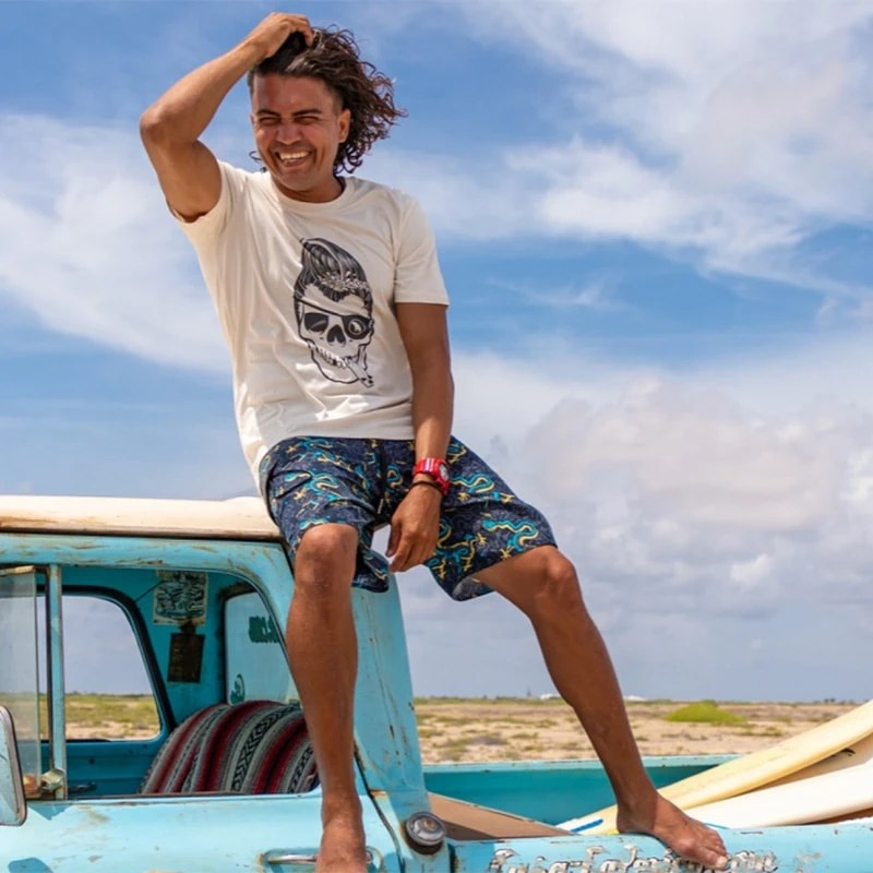 Model sits smiling on the roof of a car with his hand in his hair and wears a white Swakiko shirt with a skull with a surfboard in the corner of his mouth.